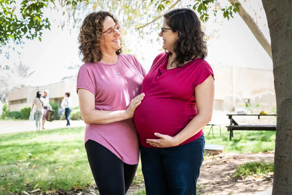 הגר בן דוד, דולה, מחייכת אל יולדת בהריון מתקדם, ידה על הבטן, בגינה מוצלת